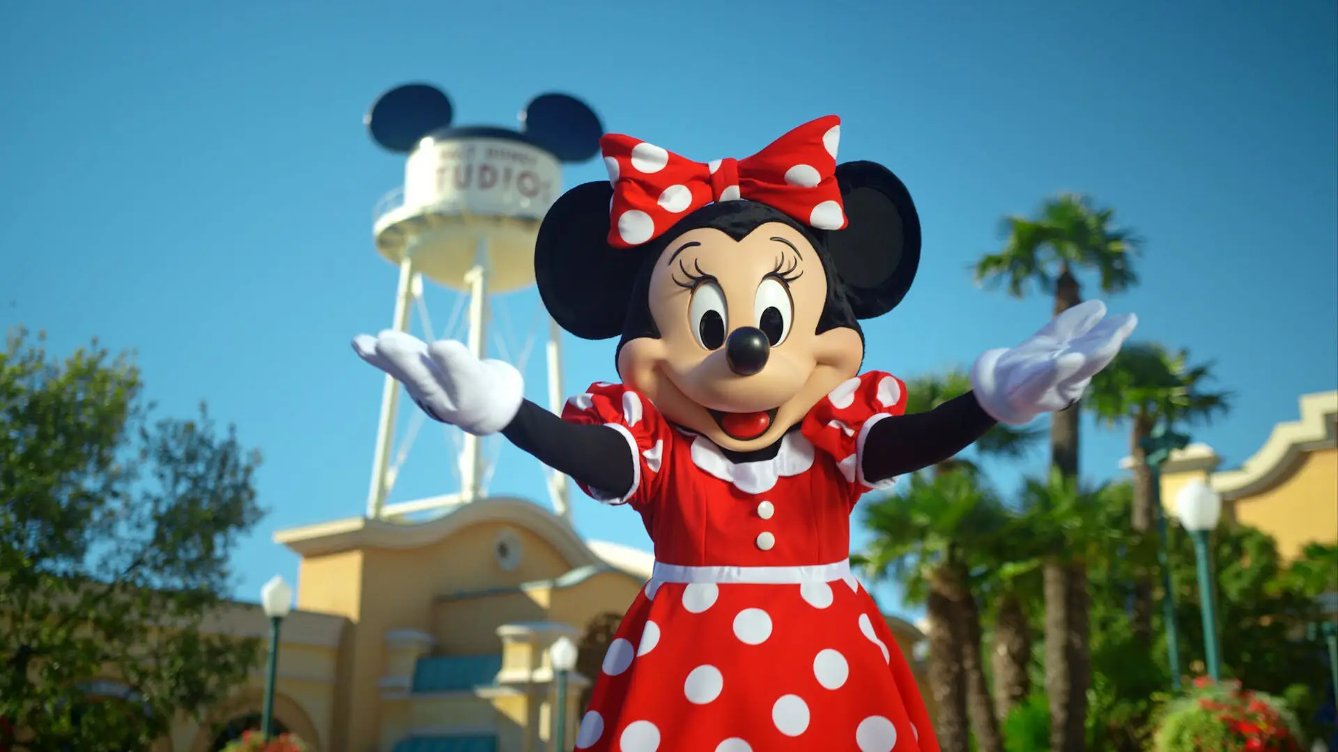 Minnie Mouse im roten Kleid mit weißen Punkten und passender Schleife lädt ein 100 Jahre Disney mitzufeiern