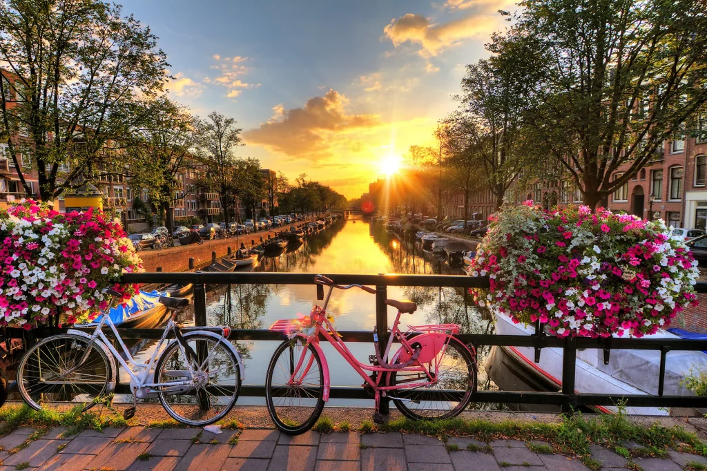 Blick von einer Brücke auf die Gracht bei Sonnenuntergang mit Geranien und bunten Fahrrädern bei Bahn und Hotel Amsterdam